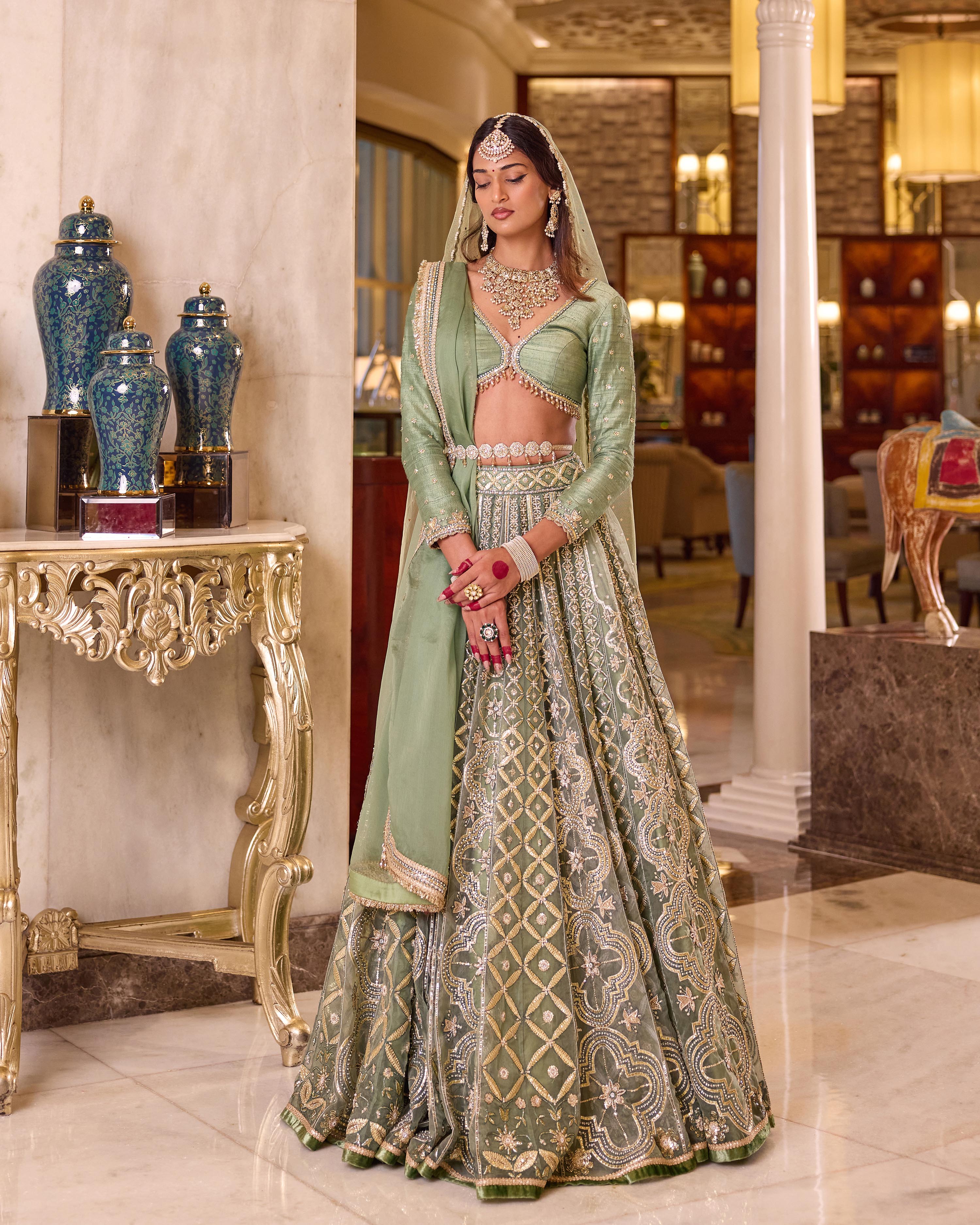 A woman models an ornate light green and gold embroidered traditional Indian lehenga choli set in an elegant indoor setting with marble floors and decorative columns.