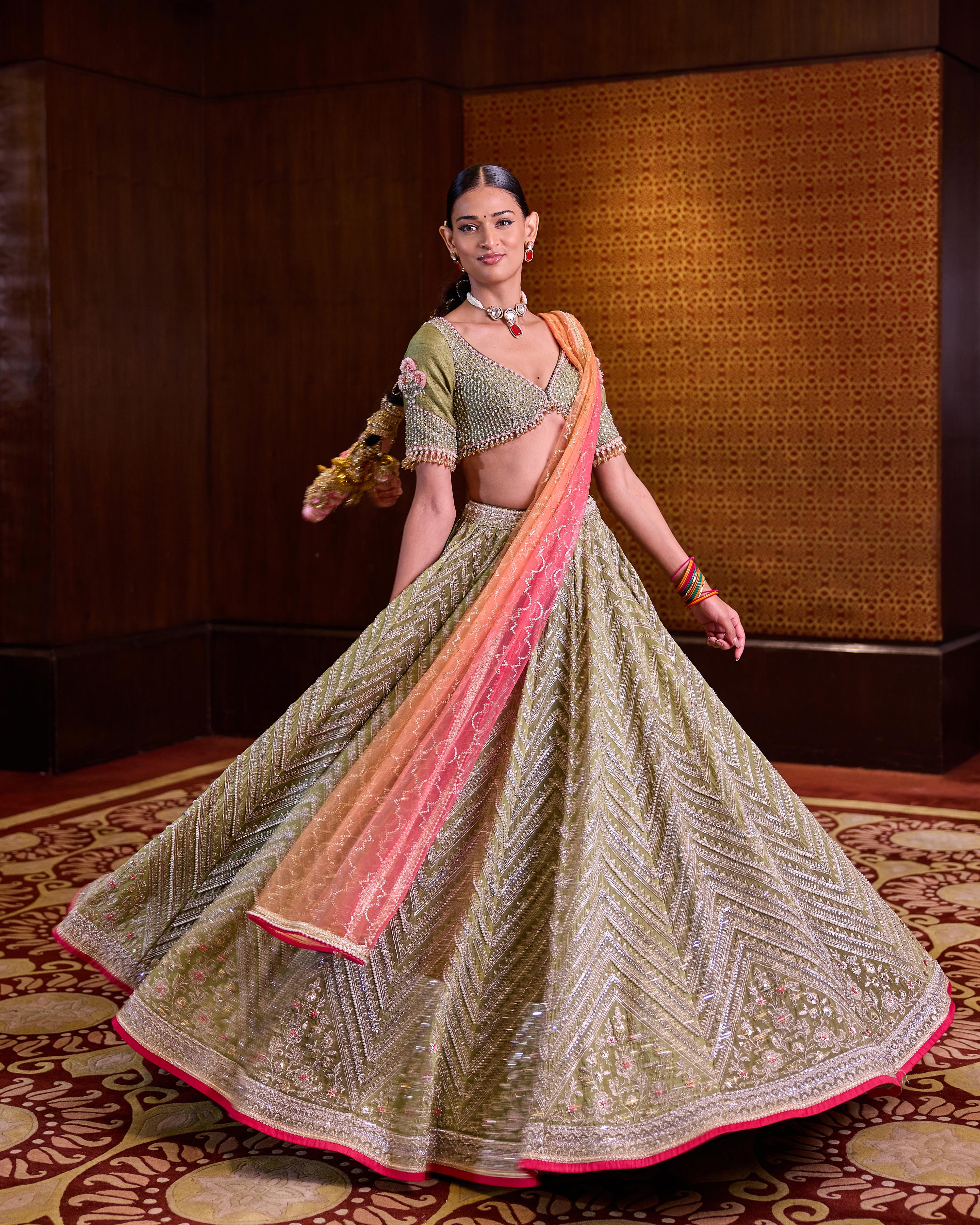 A woman models a traditional Indian lehenga choli suit. The outfit features a heavily embroidered olive green skirt with gold zari work, a matching green choli, and a peach-to-pink gradient dupatta draped over the skirt.