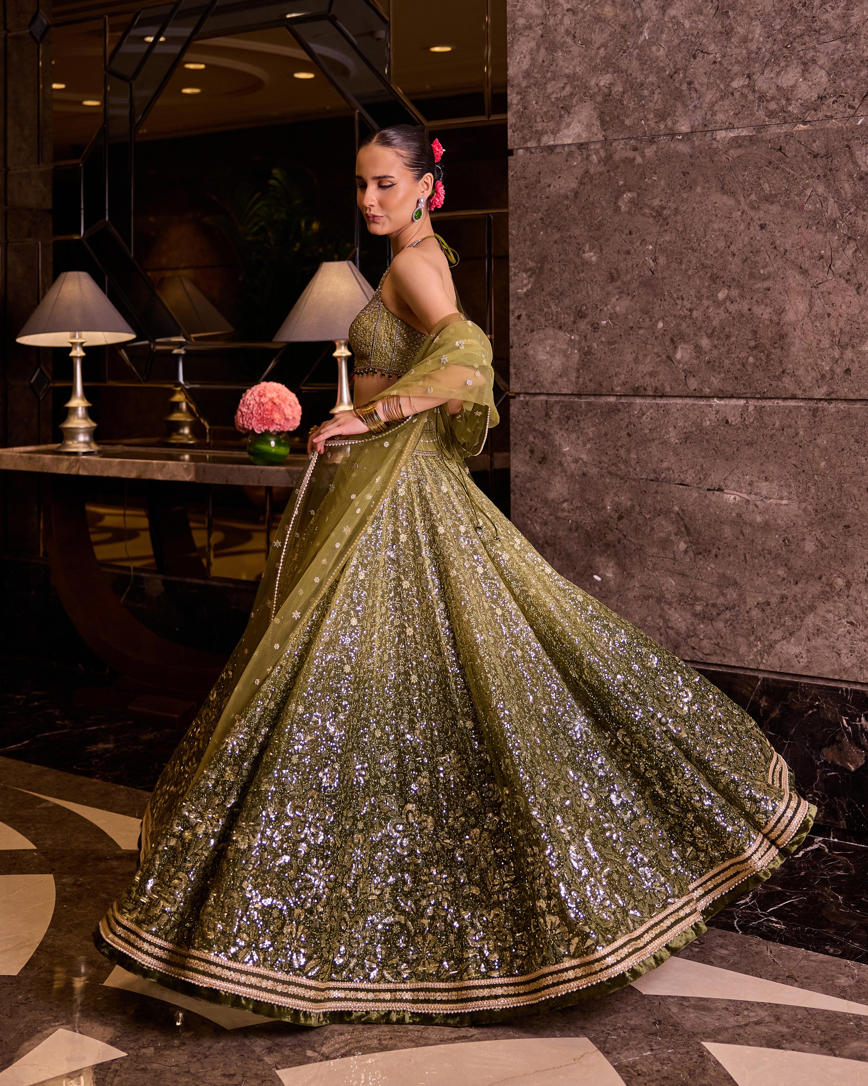 A woman models a traditional Indian lehenga choli set in olive green with intricate silver sequin embroidery, set against a dark marble interior background.